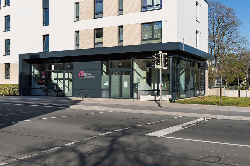 Modernes Gebäude mit großen Glasfenstern im Erdgeschoss. Die Straße und der Zebrastreifen befinden sich im Vordergrund, und im Hintergrund sind Bäume zu sehen.