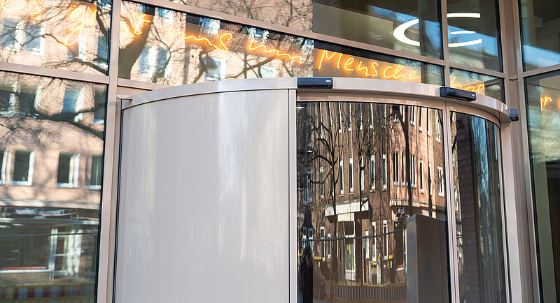 Eine moderne gläserne Drehtür mit Spiegelungen der umliegenden Gebäude und Bäume vor einer Fassade mit großen Fenstern und orangefarbener Neonschrift über dem Eingang.