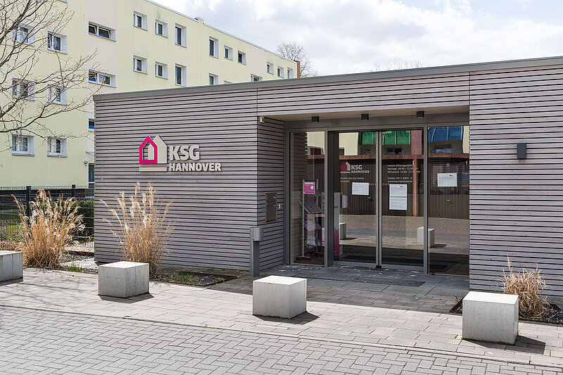 Ein modernes Gebäude mit horizontaler Holzverkleidung und Glastüren trägt das Logo und den Namen der KSG Hannover in Rosa und Weiß. An den Türen sind Aushänge angebracht, und im Hintergrund sind Wohnhäuser zu sehen.
